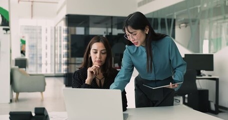 Two multi-ethnic women colleagues working together in office, engaged in collaborative discussion, reviewing and analyzing important documents or data displayed on laptop. Teamwork and productivity