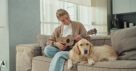 A young woman sitting on the couch, playing the ukulele next to her golden retriever, in a bright, modern living room, enjoying a relaxing moment with her pet by her side