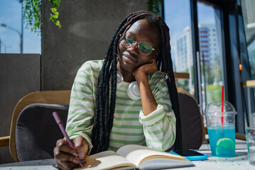 Young woman is sitting at a table in a cafe, enjoying a blue beverage and taking notes in a...