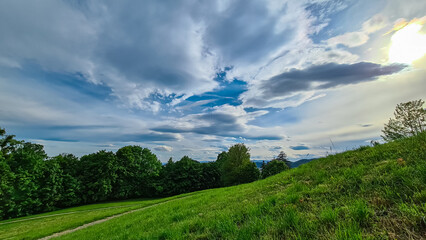 Fototapeta premium Picturesque landscape with rolling hills covered in lush green forests in Andritz, Graz, Styria. Remote woodland and meadows with backdrop of bright blue sky with dramatic white clouds. Natural beauty