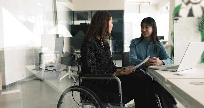 Office employee with disability sits in wheelchair lead talk with colleague at workplace, holding papers, explains contract details to workmate or company client, met in coworking space for teamwork