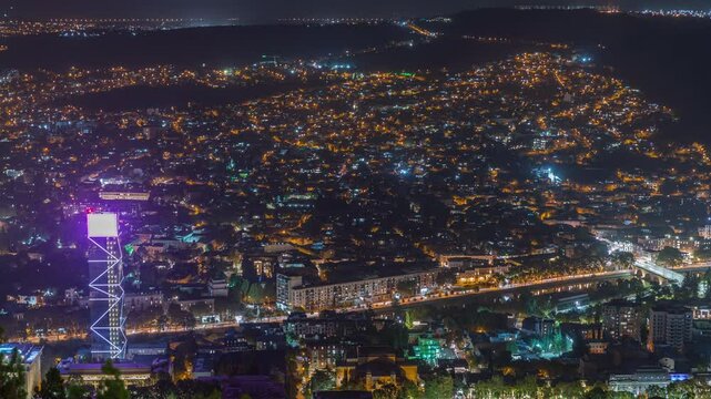 Illuminated glass tower in Tbilisi aerial timelapse, skyscrapers of Georgia, located on busy avenue. Historic building and river. View from viewpoint