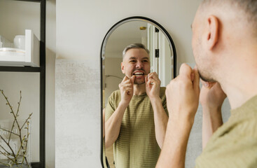 A man is flossing his teeth in the bathroom, looking intently in the mirror. His expression is focused as he carefully uses dental floss to clean between his teeth.