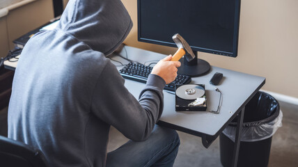 A person in a hoodie smashing a hard drive with a hammer at a desk, emphasizing digital destruction and cybersecurity risks.