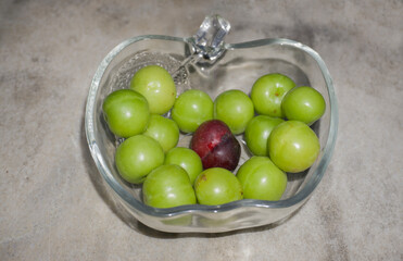 A bowl filled with green plums and a single red plum in center, highlighting contrast and individuality amidst uniformity. Evokes themes of being different, standing out, and uniqueness in a group.