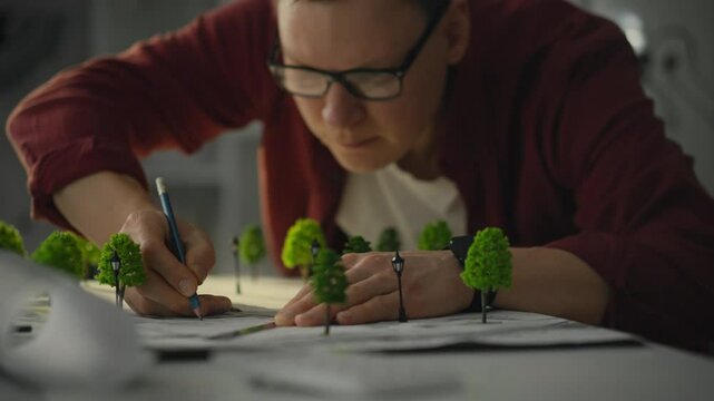 The designer focuses intently on creating an intricate architectural model, carefully positioning tiny trees while sketching plans. This demonstrates a dedication to precision and creativity