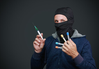 A man in a blue hoodie and black mask holding a syringe in one hand and several blood collection tubes in the other. Only his eyes are visible, giving an intense and focused look. The dark background