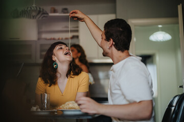 A cheerful couple shares a playful moment in a cozy kitchen while eating pasta. The scene captures joy and companionship in a relaxed home environment.