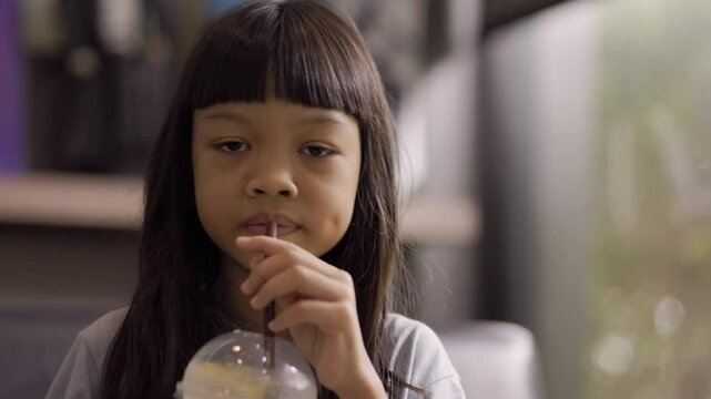 A cute Thai girl is drinking water from a brown straw. After tasting it, she makes a funny face because the drink in her glass has too much lemon sourness.