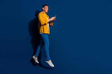 Naklejka premium Full body length photo of young guy wear jeans yellow shirt jumping with smartphone going smiling isolated on dark blue color background