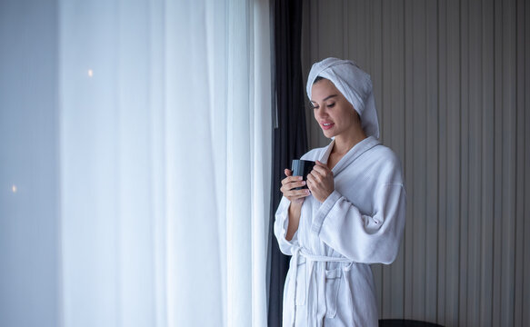 Happiness young woman in a bathrobe is smiling and feeling fresh after the shower   holding a cup of coffee while  near a window at hotel is holiday relaxing concept