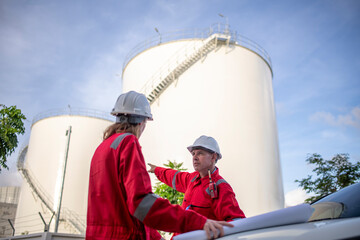 Engineers wearing safety gear, including hard hats examining survey a large blueprint tablet standing industrial facility gas or oil refinery engaged in a job requires high safety standards concept.