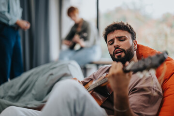 A young man strums a guitar while sitting indoors, surrounded by friends in a relaxed atmosphere. The scene conveys creativity, music, and enjoyment.