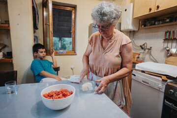 An elderly woman wraps dough while a young boy watches in a warm, homely kitchen. The scene...