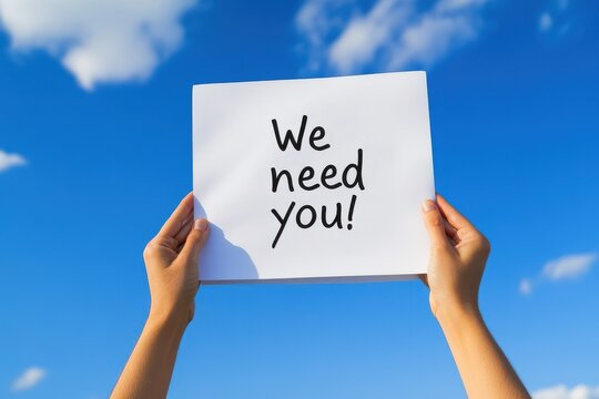 Hands holding a "We need you!" sign against a bright blue sky.
