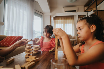 Two young girls focus on building a wooden block tower in a cozy living room, showcasing teamwork and concentration.