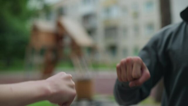 Close-up of hands interacting in a playful knuckle bump, one person in a grey top extends their hand to connect with another, with a blurred background featuring residential buildings