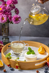 Person pouring lemon tea from teapot on wooden tray with yogurt and fruits