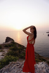 beautiful slavic woman with dark hair in luxurious red dress and accessories posing on the hill overlooking sea and sunset view