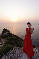 Fototapeta premium beautiful slavic woman with dark hair in luxurious red dress and accessories posing on the hill overlooking sea and sunset view