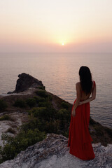 Fototapeta premium beautiful slavic woman with dark hair in luxurious red dress and accessories posing on the hill overlooking sea and sunset view