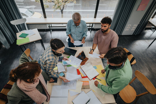 A group of college students work together on a project in a contemporary classroom, guided by an elderly professor who provides assistance and support.