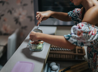 A woman in a floral dress prepares a healthy meal, carefully placing ingredients into a glass container in a modern kitchen setting.