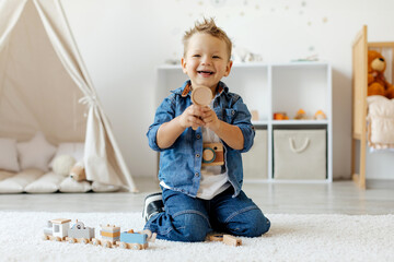 A joyful child is kneeling on the floor, holding up a wooden toy in a colorful playroom, surrounded...