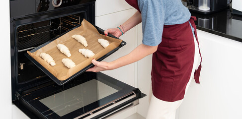 A young housewife in an apron is holding a baking sheet with croissants and is about to put them in...