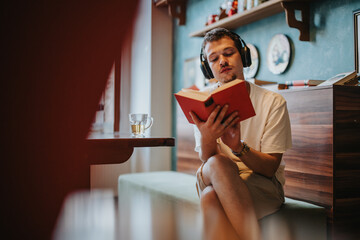 A young man wearing headphones is absorbed in reading a book indoors, creating a serene and focused...