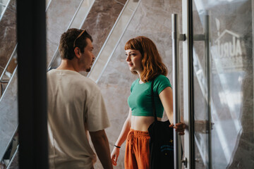 Two young adults engage in a friendly conversation indoors near a glass door. The atmosphere is casual and relaxed, highlighting everyday interactions and communication.