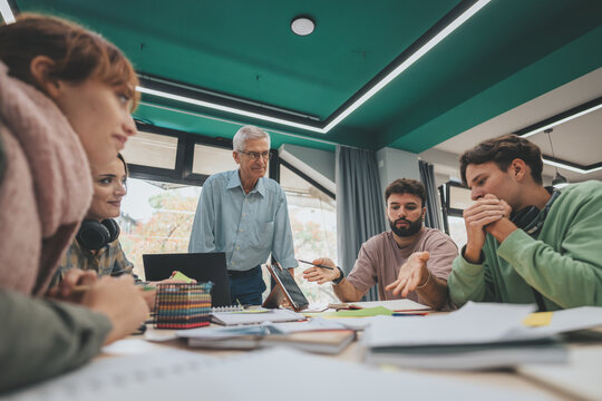 Group of university students engage in a collaborative project discussion, receiving guidance and insights from their experienced professor in a modern classroom environment.