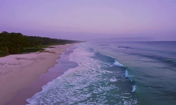Dolly shot with soft twilight: The camera starts by focusing on a quiet, secluded beach with gently rolling waves and fine, white sand. The camera moves forward, revealing the soft, purple hues of twi