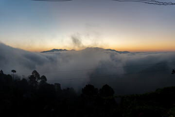 Before sunrise view, painted sky with soft orange light in the horizon. Dense clouds covers the valley and mountains in background above the clouds. Magical morning scene in mountain village in India.