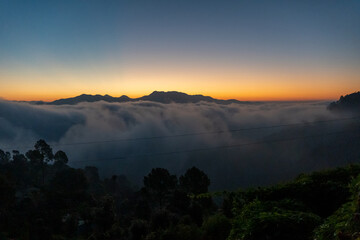 Before sunrise view, painted sky with soft orange light in the horizon. Dense clouds covers the valley and mountains in background above the clouds. Magical morning scene in mountain village in India.