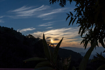 Textured clouds in orange and white hues like brush strokes on a canvas.Blue skies with textured clouds.Silhouette of layered mountains and trees creates a perfect sunset landscape in mountain village