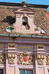A close-up view of a historic building facade in Sibiu, Romania. The facade features a pink-colored exterior with intricate decorative elements. The building has a red-tiled roof with a dormer window.