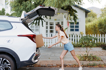 Woman unloading a cardboard box from the trunk of a white SUV car against a residential backdrop with greenery Sunny day, casual summer outfit, focused on her task