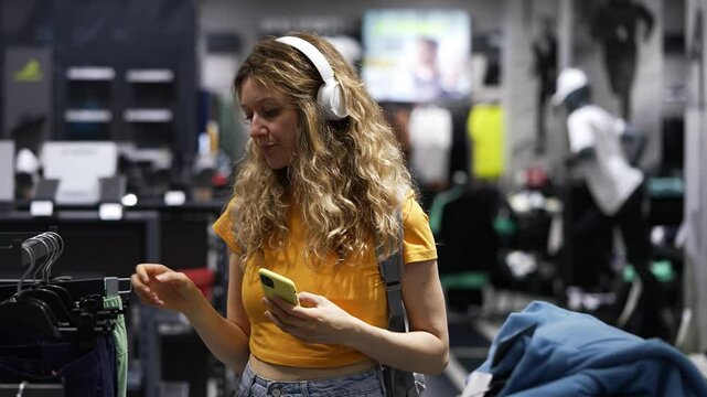 Young woman wearing headphones compares clothing prices on her smartphone while shopping in a retail store, blending technology with fashion in a modern shopping experience