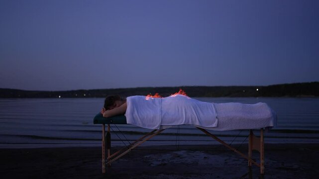 Therapist places fire cups on the back of a patient lying on a massage table by a tranquil lake at dusk, promoting relaxation and holistic healing through cupping therapy