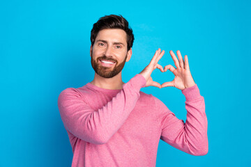 Fototapeta premium Smiling man in a pink sweater making a heart shape with his hands on a blue background, expressing love and happiness
