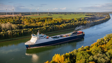 Car ferry makes its way through the Kiel Canal, bordered by  towering wind turbines, ground-mounted...