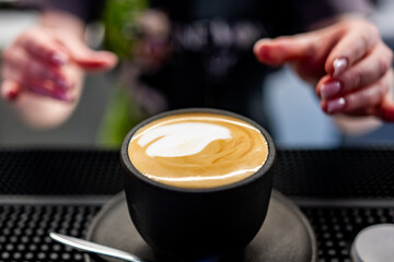 A close-up of a cappuccino with intricate latte art in a black cup on a saucer. Two hands are visible above the cup, suggesting the barista’s presentation. Ideal for culinary or lifestyle themes.