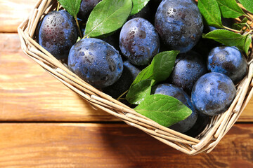 Tasty ripe plums and leaves in wicker basket on wooden table, top view