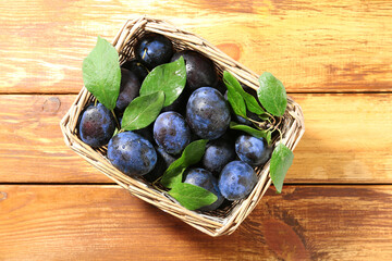 Tasty ripe plums and leaves in wicker basket on wooden table, top view