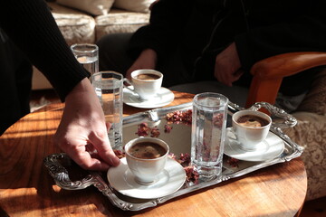 The woman taking the coffee from the silver tray and her husband sitting on the sofa in the back drinking coffee.Traditional turkish coffees and glass of waters on silver tray.Drinking coffee mutually