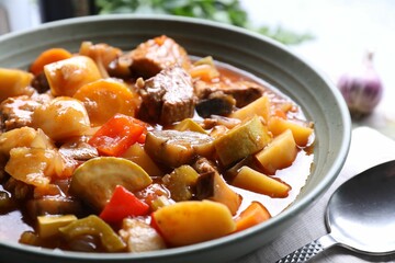 Delicious stew with vegetables in bowl and spoon on table, closeup