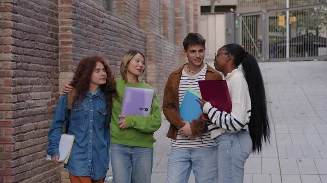 Happy students gathered together standing speaking quietly on university campus with backpacks and holding notes. Young diverse friends chatting and looking at each other, outdoors recreation people 