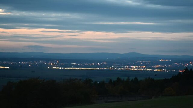 Sunset view from Buchenwald, Weimar, Germany, with a distant city, grassy fields, old stone wall, trees, and an orange-blue cloudy autumn sky.
