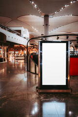 A blank, vertical advertisement display screen mockup stands in a spacious, modern shopping mall or an airport terminal The sleek, glossy interior features stylish wooden elements and ambient lighting